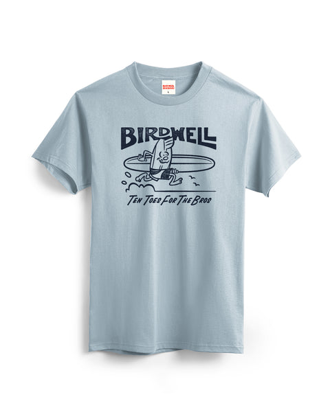 The B-Line T-Shirt - Light Blue is a garment-dyed cotton tee featuring a surfboard character with arms and legs surfing and the text “BIRDWELL TEN TOES FOR THE BROS.”.