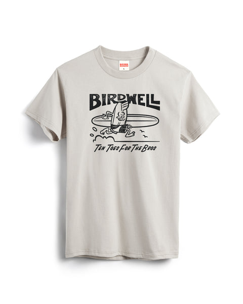 The B-Line T-Shirt - Cement is a light gray heavyweight tee featuring a cartoon surfing bird with 'BIRDWELL' above and 'TEN TOES FOR THE BROS' below. Garment-dyed for a unique, lived-in feel.