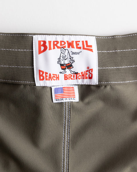 Close-up of the 808 SurfStretch Side Pocket Boardshorts - Army waistband with a white Birdwell Beach Britches label in red featuring a cartoon mascot, plus a small American flag tag reading 