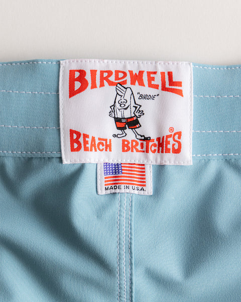 Close-up of a white Birdwell Beach Britches label stitched onto light blue 808 SurfStretch Side Pocket Boardshorts, featuring a cartoon bird mascot, red text, and a small American flag tag. Made proudly in the USA.
