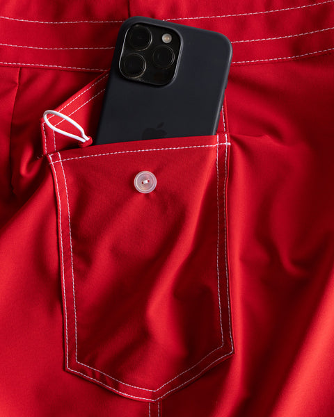 A black smartphone peeks out from the buttoned back pocket of bright red 808 SurfStretch Side Pocket Boardshorts with white stitching, made in the USA.