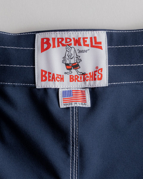 A close-up shows the Birdwell Beach Britches label with a cartoon bird on navy fabric. Below, a small tag displays an American flag and 