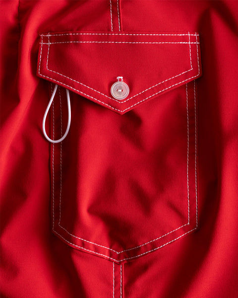 A close-up of the 806 SurfStretch Side Pocket Boardshorts in red shows white stitching and a button closure, with a white earbud wire partly out—showcasing quality USA-made craftsmanship.