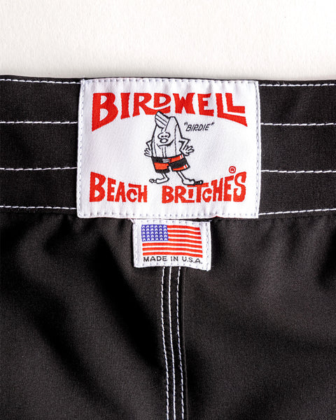 Close-up of the 806 SurfStretch Side Pocket Boardshorts - Black label on black fabric. The top tag shows Birdwell Beach Britches with a cartoon bird in shorts; below, a smaller American flag tag reads 