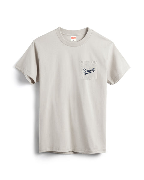 The Baller Pocket T-Shirt - Cement is a light gray, garment-dyed, heavyweight cotton short-sleeve tee with a left chest pocket featuring navy Birdwell script embroidery. Made in USA, shown on a white background.
