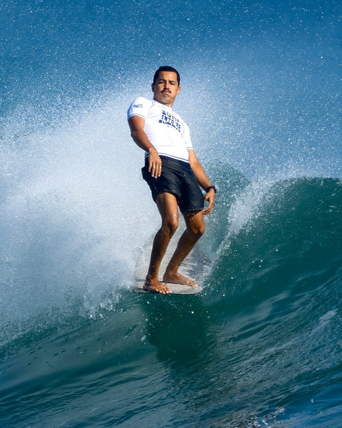 A man wearing 300 Boardshorts - Black surfs confidently on a wave, balancing as water sprays around him beneath a clear blue sky.