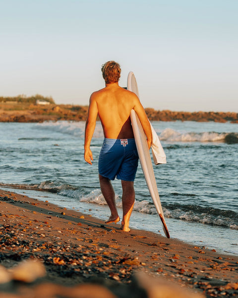 A man walks barefoot along a sandy beach, carrying a white surfboard under his arm. He is wearing 300 Boardshorts - Royal Blue. Ocean waves roll in with rocks in the background beneath a clear sky.
