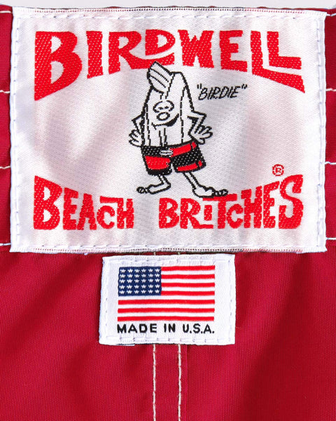 A label on the 300 Boardshorts - Red shows Birdwell Beach Britches with a cartoon bird in red shorts, above a Made in U.S.A. tag with an American flag—classic boardshorts made from red SurfNyl fabric.