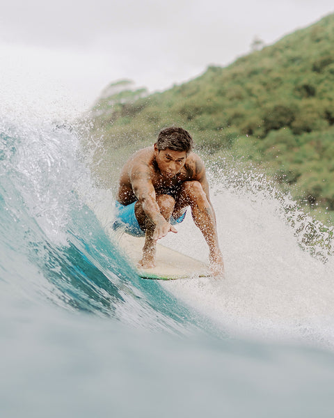 A man in red 300 Boardshorts crouches low on a surfboard, riding a wave near a green, tree-covered hill under a cloudy sky. Water splashes around him as he concentrates on keeping his balance.