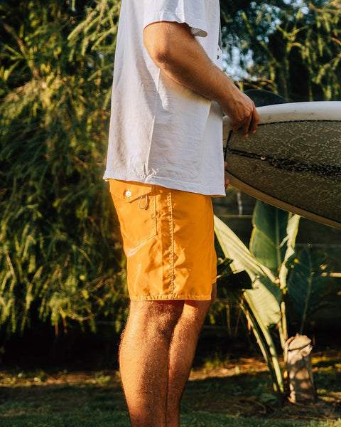 Wearing 300 Boardshorts - Gold, a person stands outdoors holding a surfboard, surrounded by green foliage and sunlight.