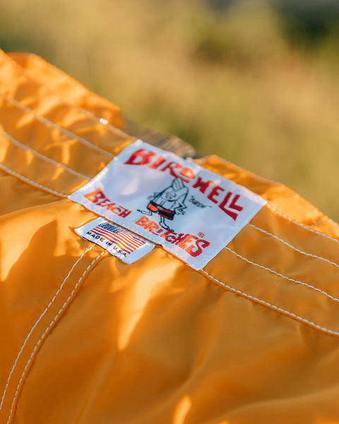 Close-up of the 300 Boardshorts - Gold, featuring a white Birdwell Beach Britches label with a cartoon bird and American flag 