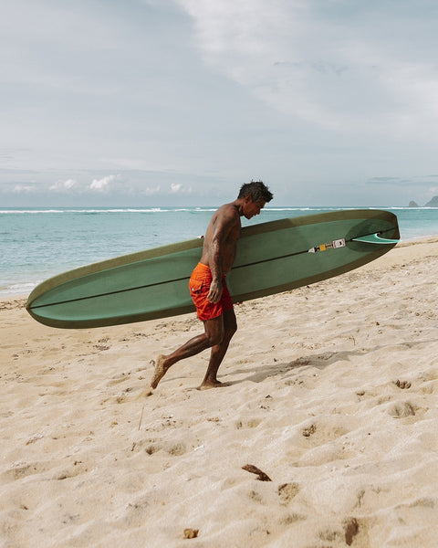 Wearing 300 Boardshorts in Medium Orange, a person carries a green surfboard under one arm while walking barefoot along the sandy beach by the ocean, with waves rolling in beneath a cloudy sky.