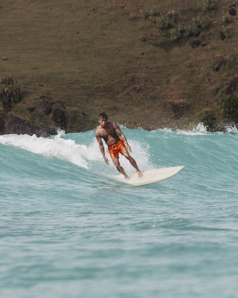 A man in 300 Boardshorts - Medium Orange surfs a light blue wave near a grassy shore, balancing on his board with a relaxed posture.
