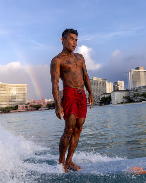 A tattooed man in navy 310 Boardshorts rides a surfboard on calm ocean water near a city skyline, with buildings and a rainbow visible in the background under a partly cloudy sky.