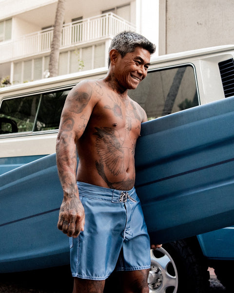 A smiling, tattooed man with gray hair stands shirtless in 310 Boardshorts - Federal Blue, holding a blue surfboard by a van. An apartment building and palm tree are visible in the background.