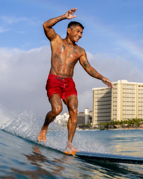 A tattooed man surfs barefoot in 310 Boardshorts - Red, with a hotel and blue sky behind him, highlighting classic surf swimwear style.