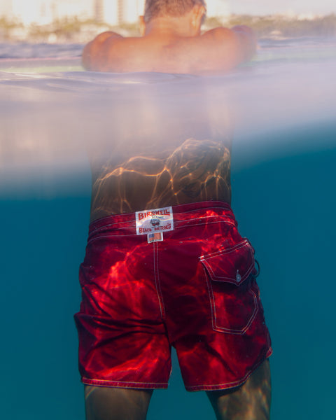 Wearing bright red 310 Boardshorts, a person stands waist-deep in clear blue water as sunlight shimmers on the fabric and their lower back. The background is softly blurred.