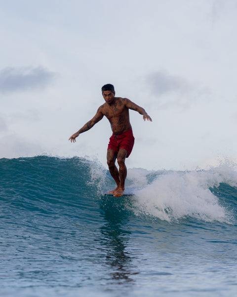 A tattooed man wearing red 310 Boardshorts is surfing a small wave on his board beneath cloudy skies.