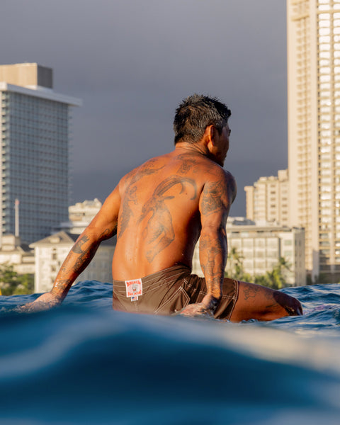 A tattooed man with short dark hair sits on a surfboard in the ocean, facing away, wearing 310 Boardshorts - Tobacco, with tall city buildings and a cloudy sky in the background.
