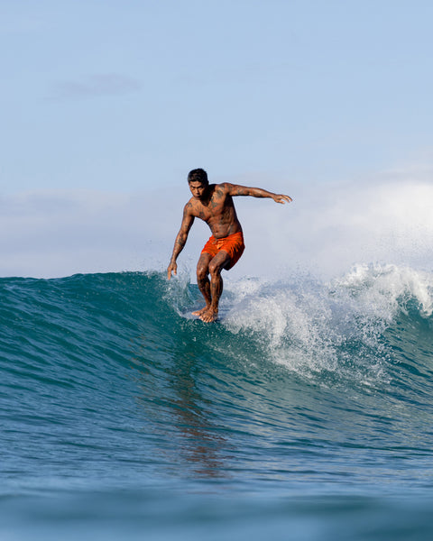 A man wearing 310 Boardshorts in Medium Orange surfs a blue ocean wave under a clear sky, balancing on his surfboard with arms outstretched.