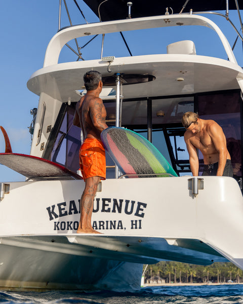 Two men in 310 Boardshorts - Medium Orange stand on the back of a boat called KEA’ENUE at Koko Marina, HI. One holds a colorful surfboard while the other leans forward, smiling under a sunny sky above clear blue water.
