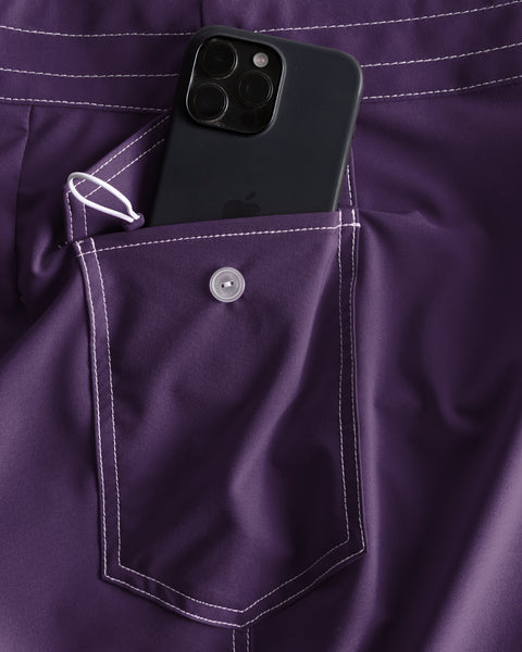 A black smartphone peeks out of the side pocket of a buttoned purple 806 SurfStretch Side Pocket Boardshorts with white stitching.