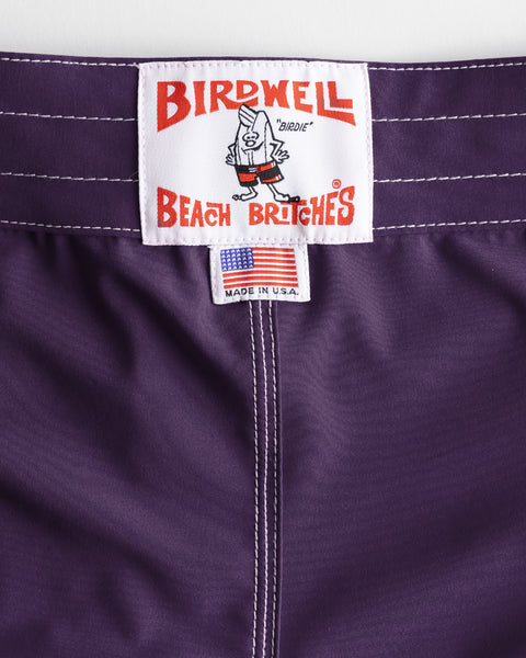 Close-up of the 808 SurfStretch Side Pocket Boardshorts in purple, featuring a Birdwell Beach Britches label with a cartoon bird, a 
