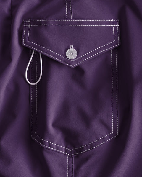 Close-up of the 808 SurfStretch Side Pocket Boardshorts in purple: features a fabric pocket with a flap, button closure, white contrast stitching, and a small white loop near the opening. Made in USA.