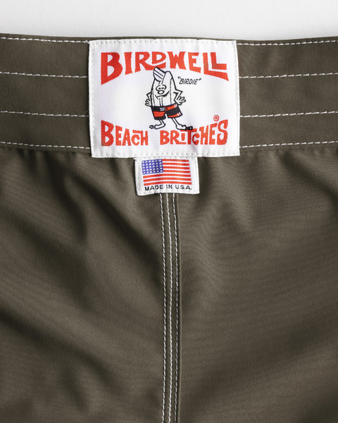 Close-up of the 806 SurfStretch Side Pocket Boardshorts - Army waistband with white stitching, showcasing a Birdwell Beach Britches label with a cartoon bird and a small tag featuring an American flag and “Made in U.S.A.” text.