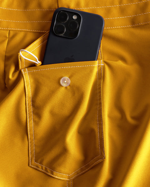A black smartphone is partially tucked into the buttoned back pocket of 806 SurfStretch Side Pocket Boardshorts - Gold, which feature white stitching and a small loop near the pocket.