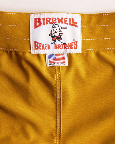 Close-up of gold 808 SurfStretch Side Pocket Boardshorts by Birdwell Beach Britches, featuring a cartoon label, white stitching, and a small MADE IN USA flag tag—highlighting their signature SurfStretch material.
