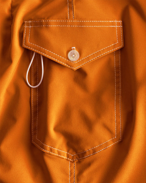 Close-up of the 808 SurfStretch Side Pocket Boardshorts in Medium Orange, showing the pocket with white stitching, buttoned flap, and a white earphone wire partially tucked inside.