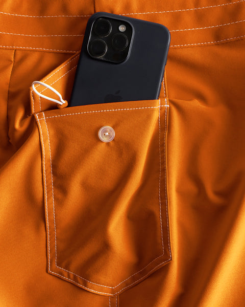 A black smartphone partially tucked into the back pocket of 808 SurfStretch Side Pocket Boardshorts in Medium Orange with white stitching.