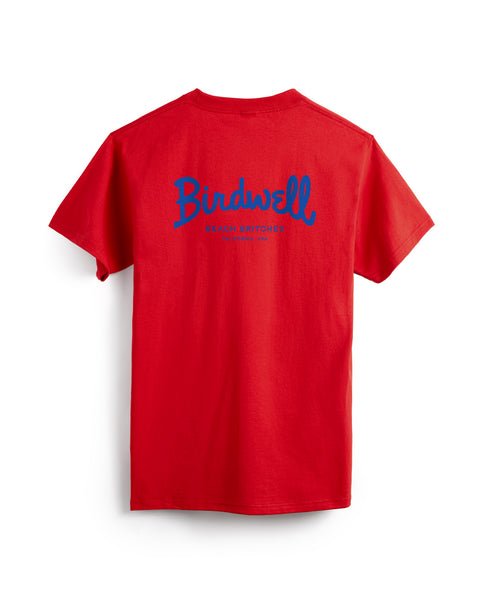The Script Pocket T-Shirt - Tomato is a red, heavyweight garment-dyed tee with large blue Birdwell script and smaller BEACH BRITCHES text on the back. The short-sleeve shirt has a pocket and is shown laid flat, back facing up.