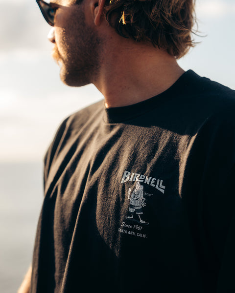 A person wearing the black 61 T-Shirt by Birdwell, made of heavyweight 100% cotton, stands outdoors. The garment-dyed tee features a small cartoon bird logo and 