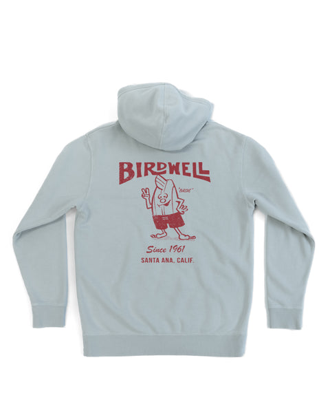 The 61 Midweight Hoodie - Sage is a light gray relaxed fit hoodie with a red screen-print of a cartoon surfboard character waving on the back, and text that reads “BIRDWELL,” “Since 1961,” and “Santa Ana, Calif.”.
