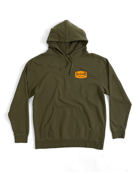 The Herald Midweight Hoodie - Army is an olive green relaxed fit hoodie with a front pocket, adjustable drawstrings, and a small yellow patch on the left chest reading 