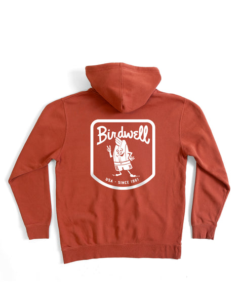 The Herald Midweight Hoodie - Paprika features a brick-red color, relaxed fit, and a large white screen print on the back with Birdwell, a cartoon bird, and 
