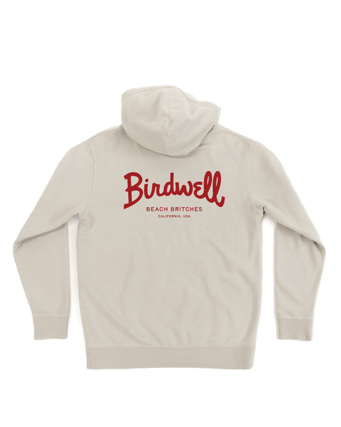 The Script Midweight Hoodie - Bone is a light beige, relaxed fit hoodie shown from the back, featuring bold red Birdwell Beach Britches California, USA text on the upper back for a classic laid-back style.
