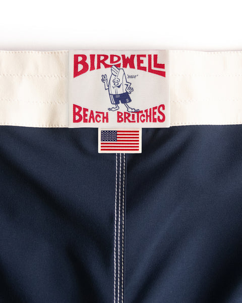 Close-up of the Birdwell Beach Britches 808 Mono Stripe - Navy/Bone label stitched onto navy fabric, showing a cartoon bird mascot and an American flag patch—a tribute to their classic USA-made boardshorts.