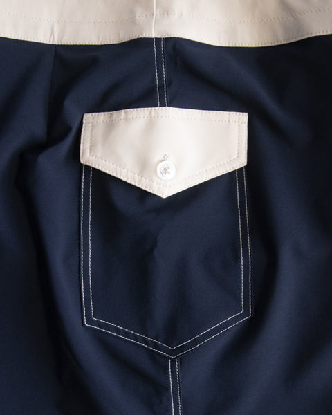 Close-up of the 808 Mono Stripe - Navy/Bone boardshort pocket featuring a triangular flap, white stitching, and white button. The top section and flap are bone to match. Made in the USA.