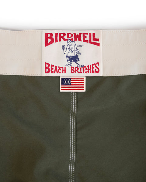 Close-up of the 808 Mono Stripe - Army/Bone boardshorts, featuring an olive green SurfStretch fabric, white waistband, signature Birdwell Beach Britches patch with cartoon bird logo, small embroidered American flag below—expertly made in USA.