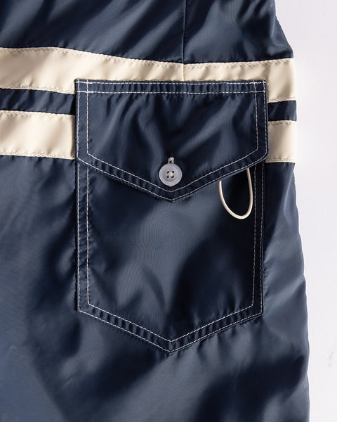 Close-up of the 300 Comp Stripe - Navy/Bone boardshorts pocket with white stitching, a flap secured by a white button, and white stripes above. A small white loop is attached at the side. Proudly made in the USA.