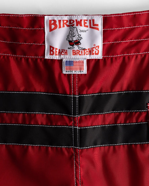 Close-up of the waistband of the 300 Comp Stripe - Red/Black boardshorts, showing two black stripes, a white Birdwell Beach Britches label with cartoon bird, an American flag 