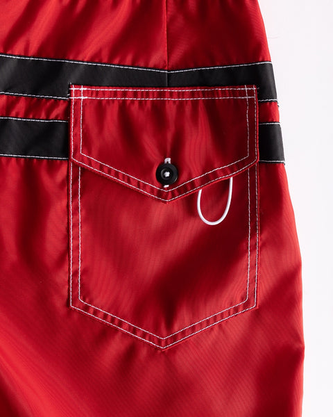 Close-up of the 300 Comp Stripe - Red/Black boardshorts pocket featuring white stitching, a black button, and white loop detail. Two black stripes cross the top; proudly made in the USA.