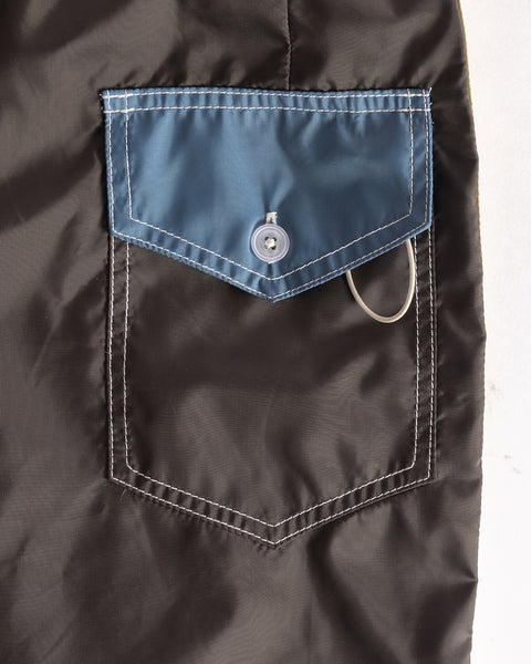 A close-up of the 300 Legend boardshorts in Brown/Federal Blue/Bone shows a pocket with white stitching and a blue flap secured by a button, partially concealing a metal keyring—crafted quality made in the USA.