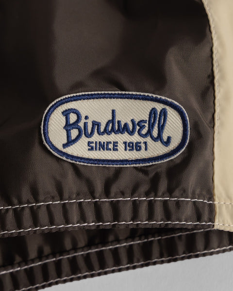 Close-up of a brown fabric patch with blue and white embroidery reading Birdwell SINCE 1961, sewn onto 300 Legend - Brown/Federal Blue/Bone SurfNyl boardshorts with elastic seam and beige trim, proudly made in the USA.
