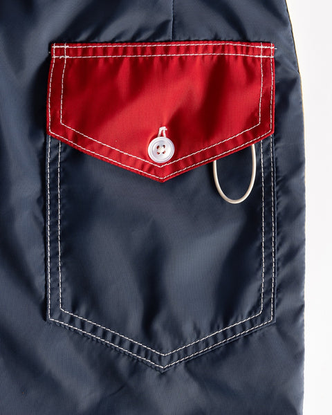 A close-up of the 300 Legend - Navy/Red/Bone boardshorts pocket shows white stitching, a red flap with white button, and a gray loop on the right. Proudly made in USA.