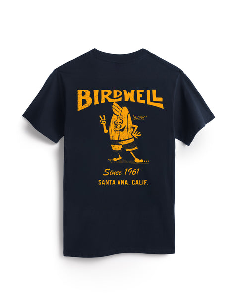 The 61 T-Shirt - Navy/Gold is a navy heavyweight cotton tee featuring a yellow back graphic of a cartoon bird in boardshorts with a surfboard and peace sign, plus “BIRDWELL,” “Since 1961,” and “Santa Ana, Calif.” text.