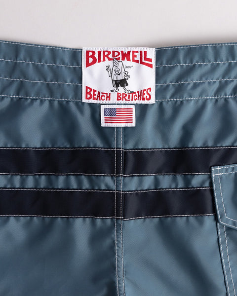 Close-up of the waistband on 300 Comp Stripe - Federal Blue/Navy boardshorts. The Birdwell Beach Britches label and an American flag patch appear above durable SurfNyl fabric with white stitching, made proudly in the USA.